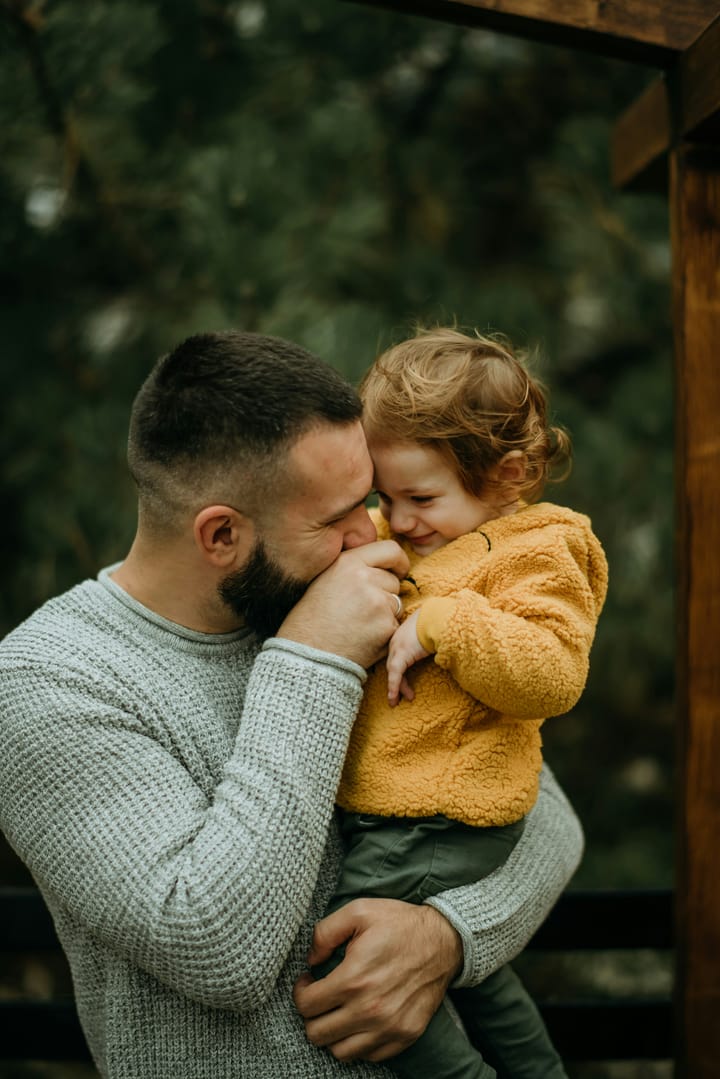 Bearded father and small child face-to-face, the father kissing the child’s hand while the child rests a hand on his cheek, expressing warmth and emotional closeness.