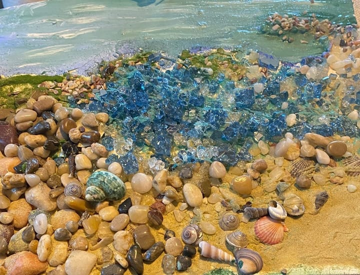 Blue-green glass fragments arranged along a sandy shoreline with small pebbles and seashells, forming a textured coastal scene.